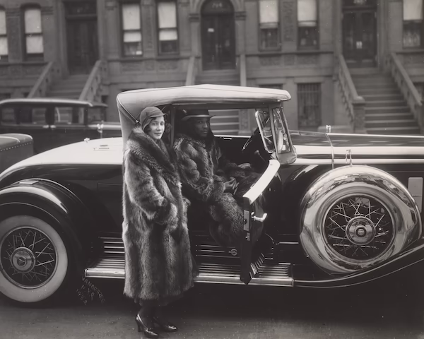 Two people in fur coats stand beside a vintage car, with one person in the driver's seat and the other standing outside. The background shows a row of brownstone buildings with stoops.